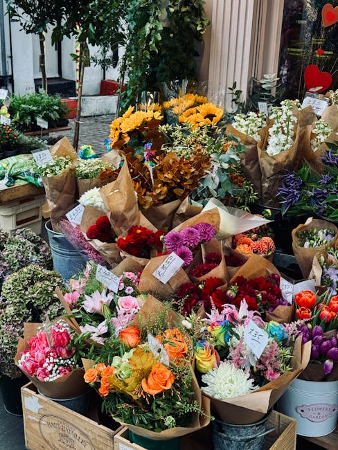 A vibrant display of numerous floral arrangements and bouquets placed outdoors on a sidewalk, arranged in metal buckets and wooden crates. The flowers include bright yellow sunflowers, deep red roses,