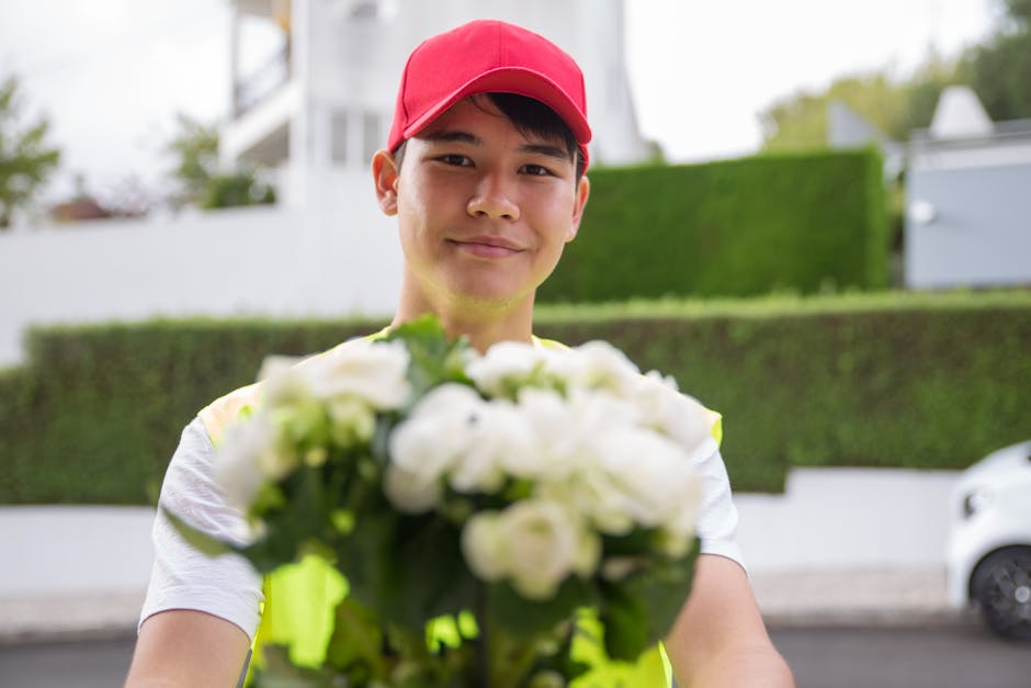 A delivery person wearing a bright red cap and a yellow high-visibility vest is smiling and holding a bouquet of fresh white flowers with green leaves, arranged in a dense cluster. The flowers are lik
