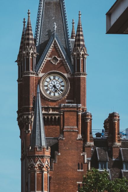 A close-up view of a tall, historic brick clock tower with pointed spires and decorative Gothic architectural details, set against a clear blue sky. The clock face displays Roman numerals and ornate c